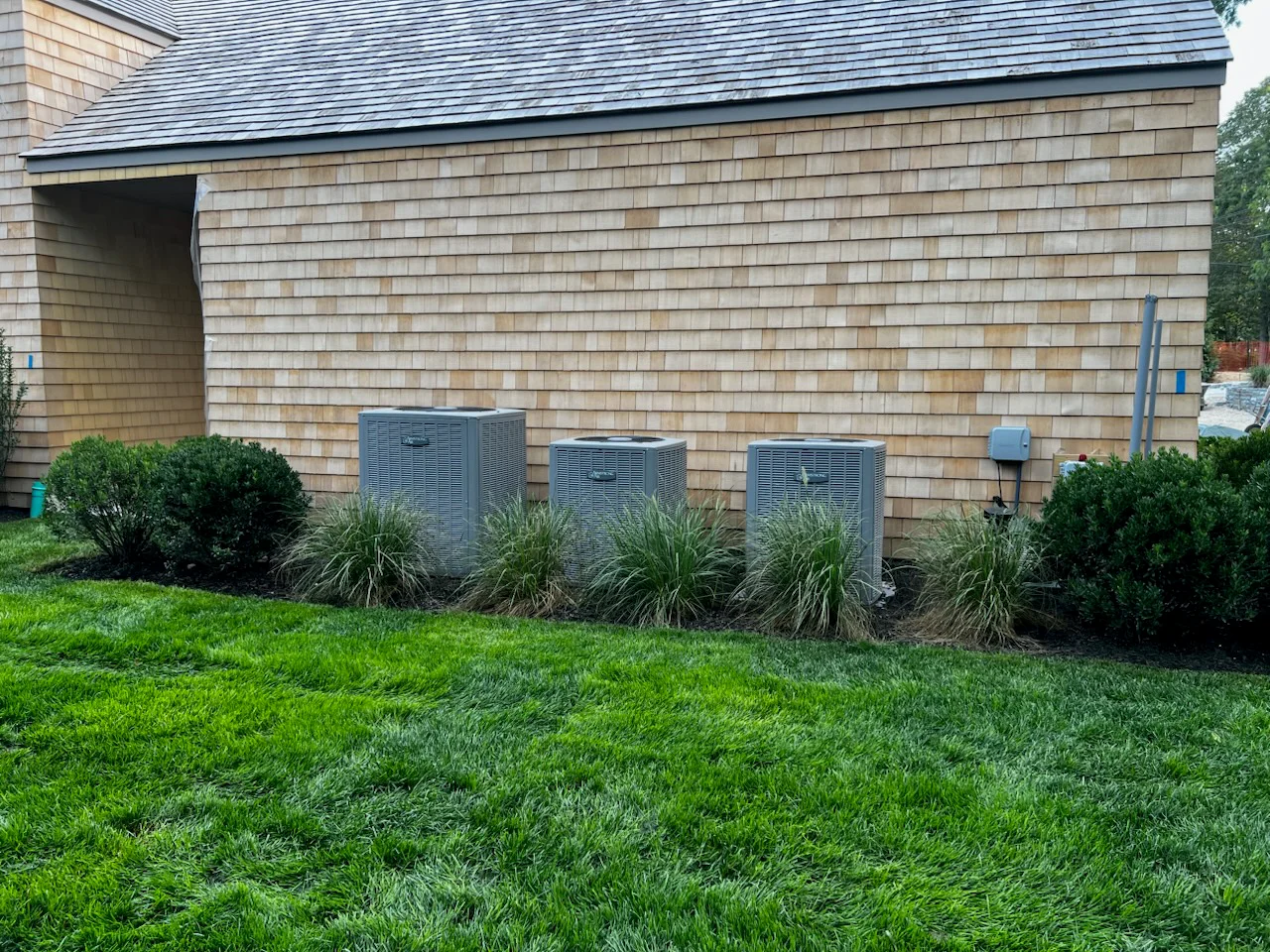 Three blue HVAC units screened with louvered covers and surrounded by ornamental grasses against cedar shake home